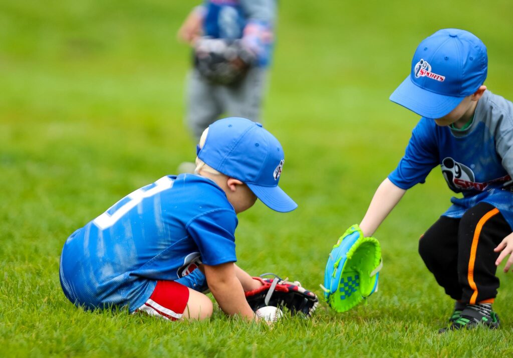 Two young kids in royal blue i9 Sports hats and jerseys kneeling on the ground with their baseball gloves to pick up a t-ball from the grass.