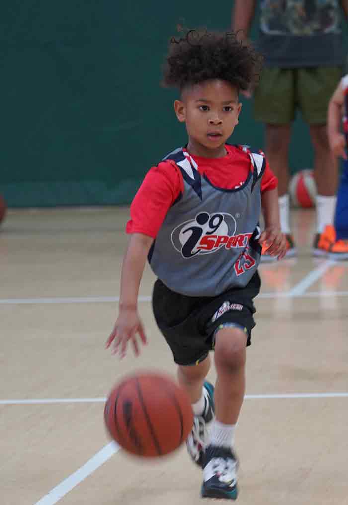 Young girl playing basketball in a gray i9 Sports basketball jersey. She is running towards you dribbling a basketball.