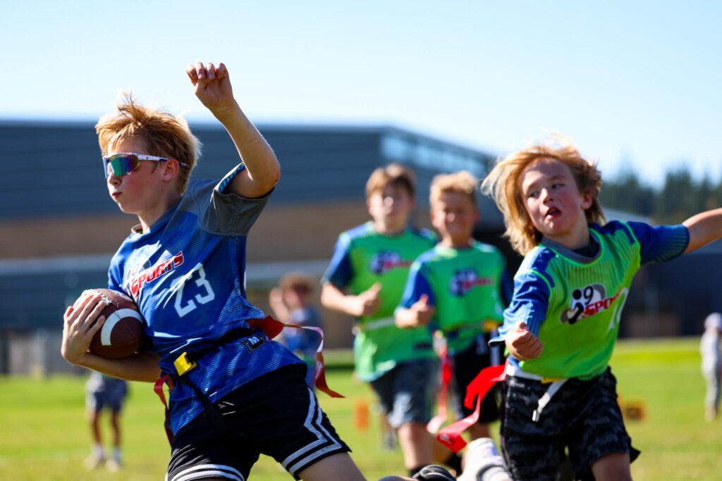 Young athletes playing flag football. On the left is an athlete running with the football in his arms. He is wearing a bright blue i9 Sprots jersey with the number 23 on it and white sport styled sunglasses. Behind him is a player of the opposite team in a bright green i9 Sports jersey attempting to pull his flag, but missing!