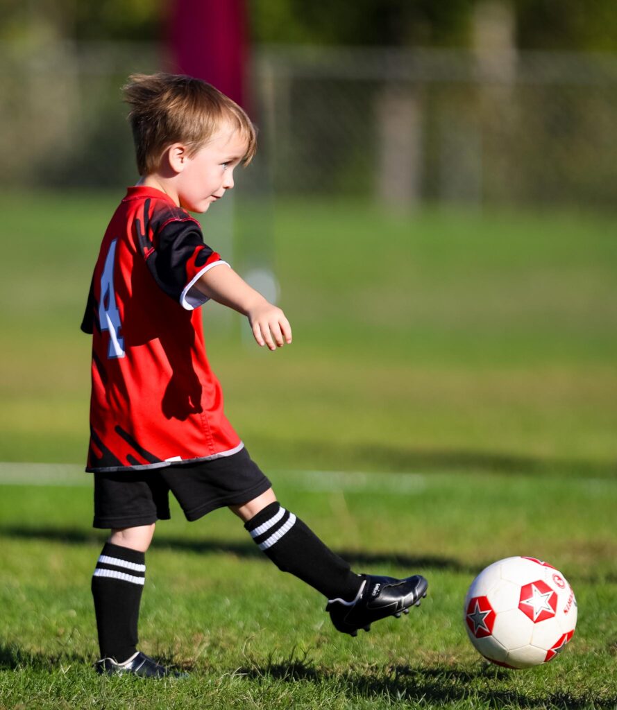 Young child in a red i9 Sports jersey kicking a soccer ball.