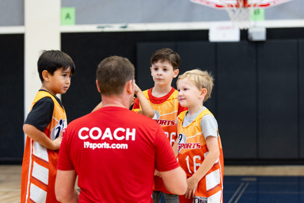 A youth basketball team in a huddle. The kids are wearing bright orange i9 Sports jerseys and the Coach has a red shirt that says COACH on the back.