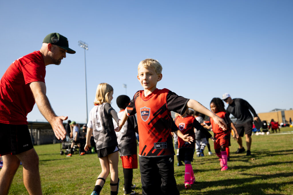 Young blond boy with red i9 Sports soccer jersey on. He is walking in line with his team after the game high giving the other team saying "good game" as a sign of sportsmanship.