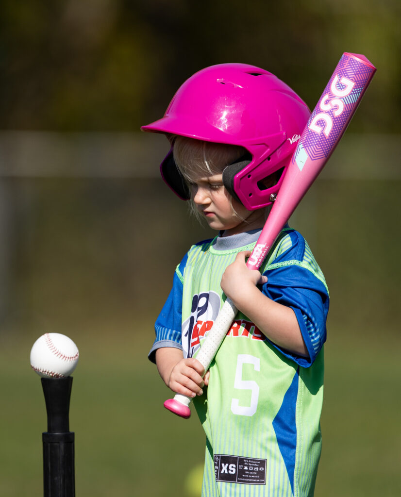 Young girl, about 4 years old, holding a bright pink t-ball pat and wearing a bright pink helmet with a blue and green i9 Sports t-ball jersey. She is standing next to a tee with a t-ball on it. The bat is sitting on her left shoulder (your right visually) waiting to hit the ball.