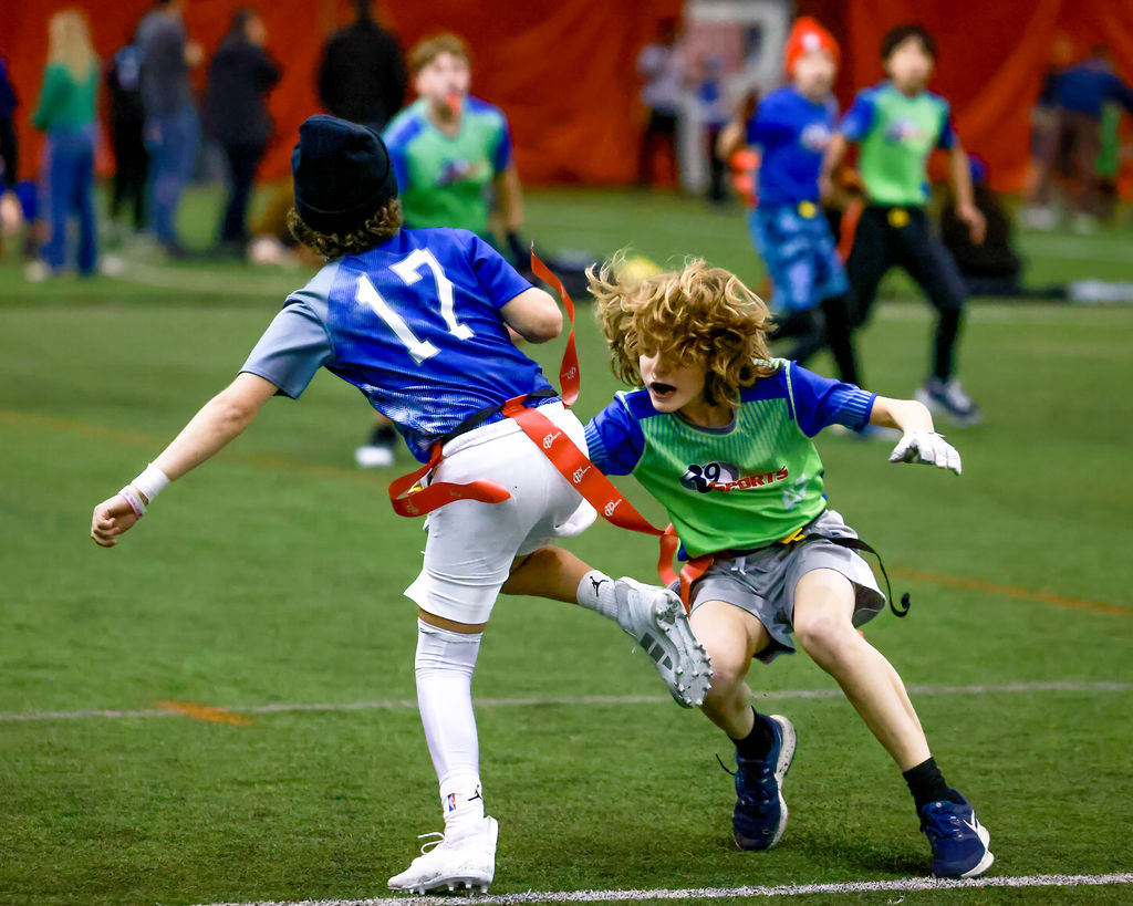 Two older kids playing flag football in bright blue and green i9 Sports jerseys. The one on your right is going for the other player on your left's flag. It's unclear in the photo if the player's flag is grabbed. Both players look very agile.