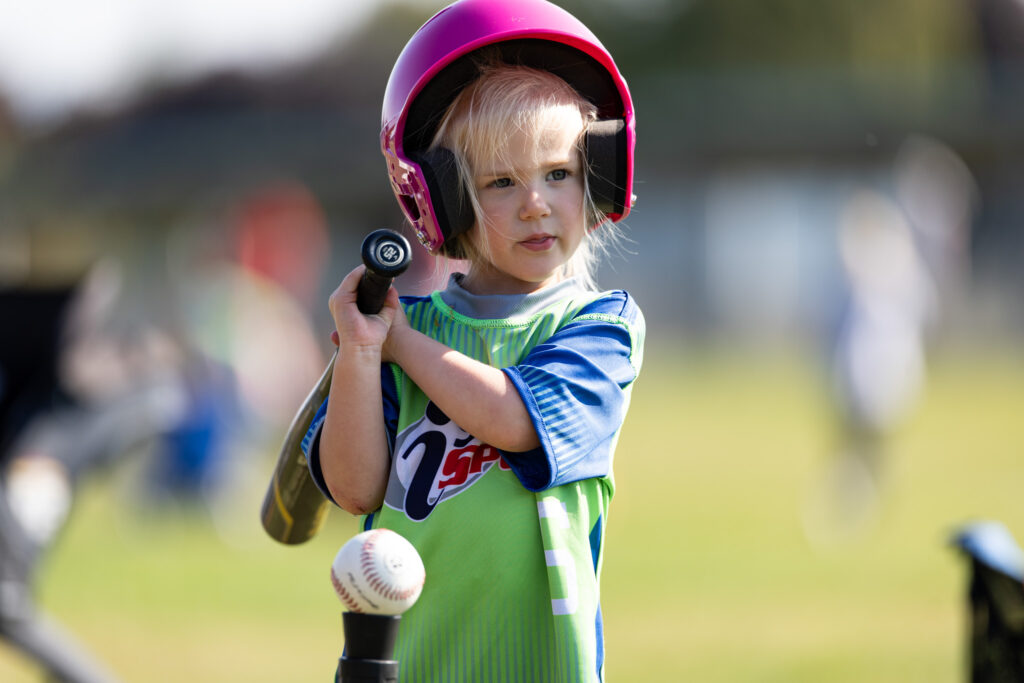 A young girl at t-ball with a pink t-ball helmet holding a bat that's too heavy for her on her right shoulder (your left) while she stands next to a ball on a tee.