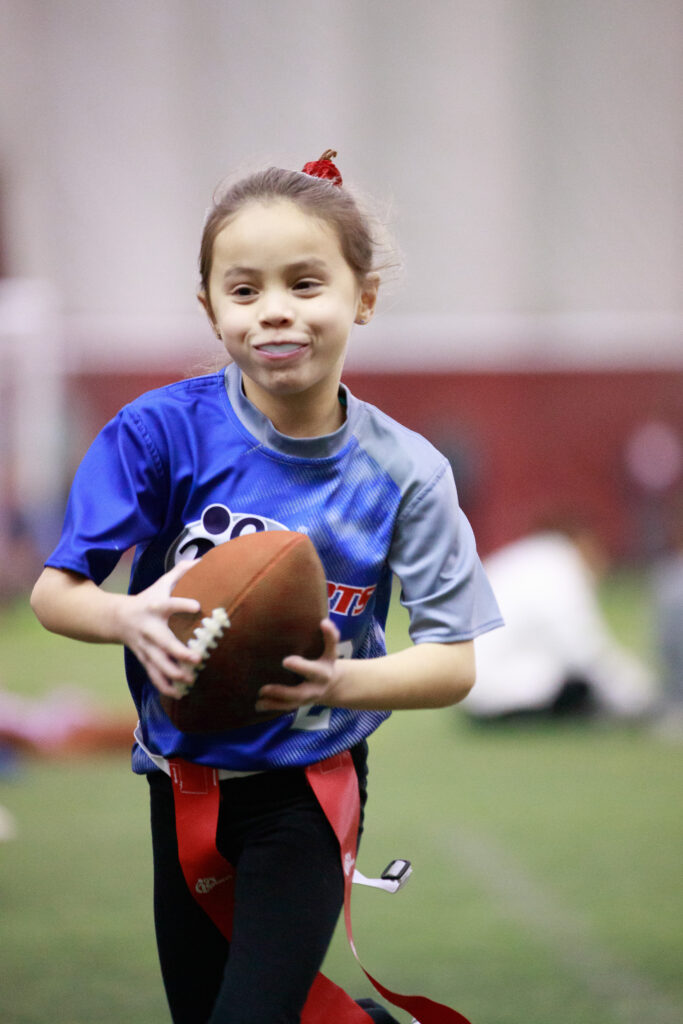 Young girl in a bright blue i9 Sports jersey playing flag football with a giant smile on her face and bright red bow in her hair that matches the red flags around her waist.