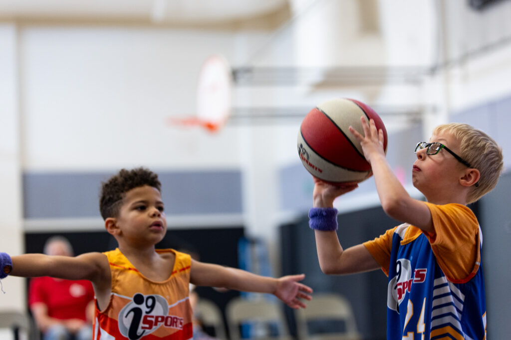 Two young boys around 5-6 years old playing basketball. The boy in the blue jersey on the right is shooting the ball while the boy in an orange jersey on the left has his arms in the air trying to block him.