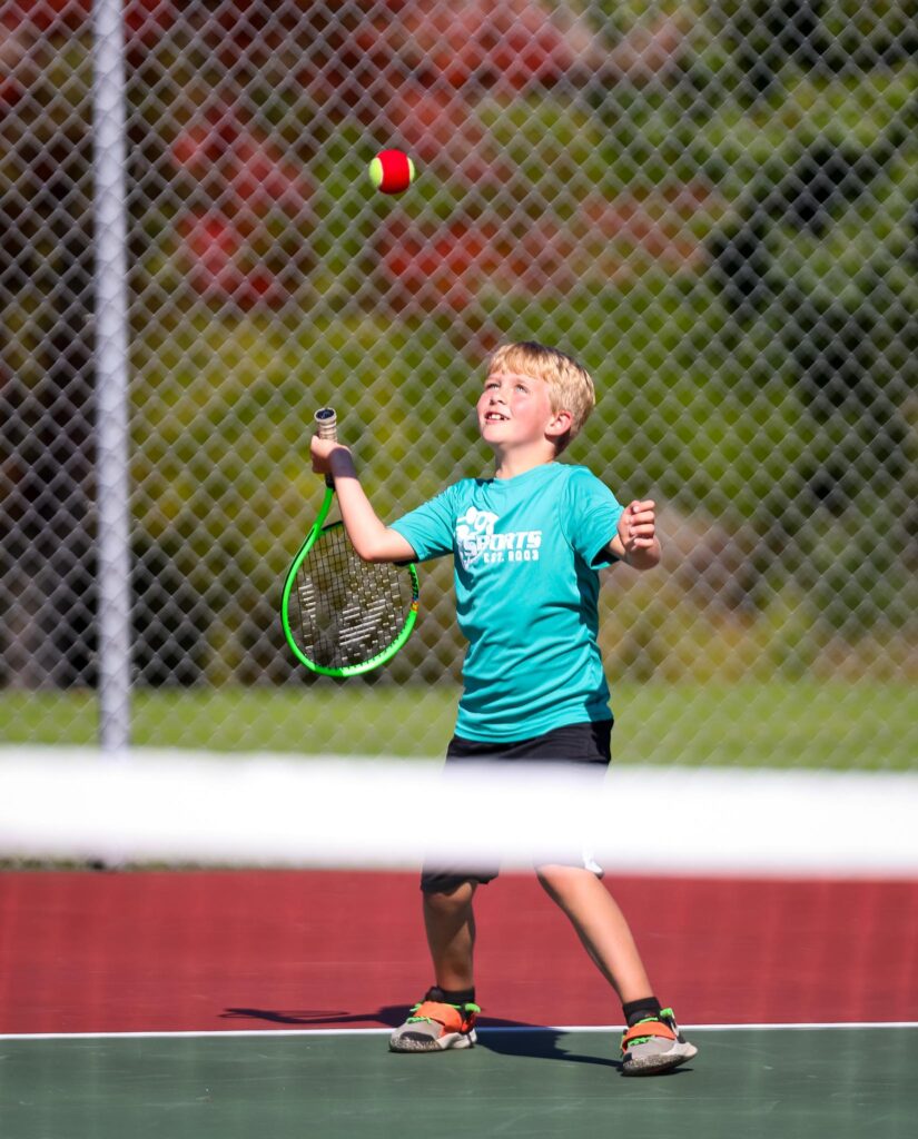 A young blond-haired tennis player practicing his overhand serve with a red and green ball.