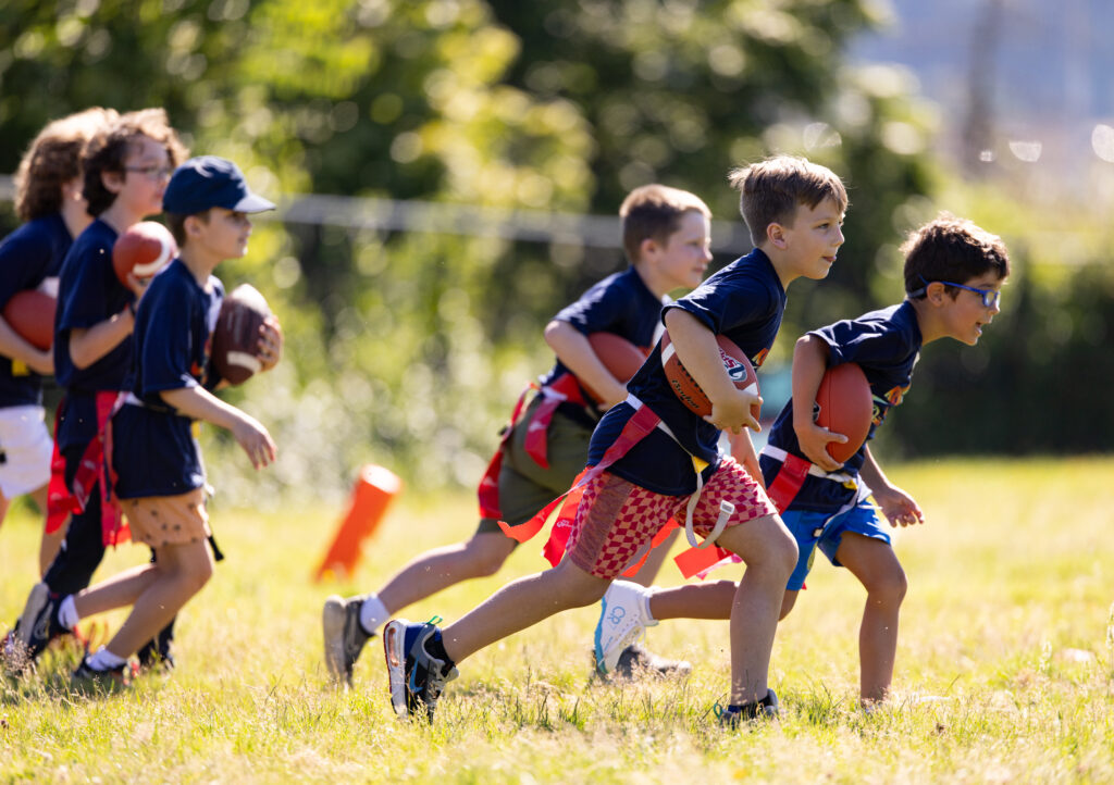 A handful of young kids at a youth sports summer camp. They are holding footballs and running while learning how to play flag football and how to cradle the ball in their arm.
