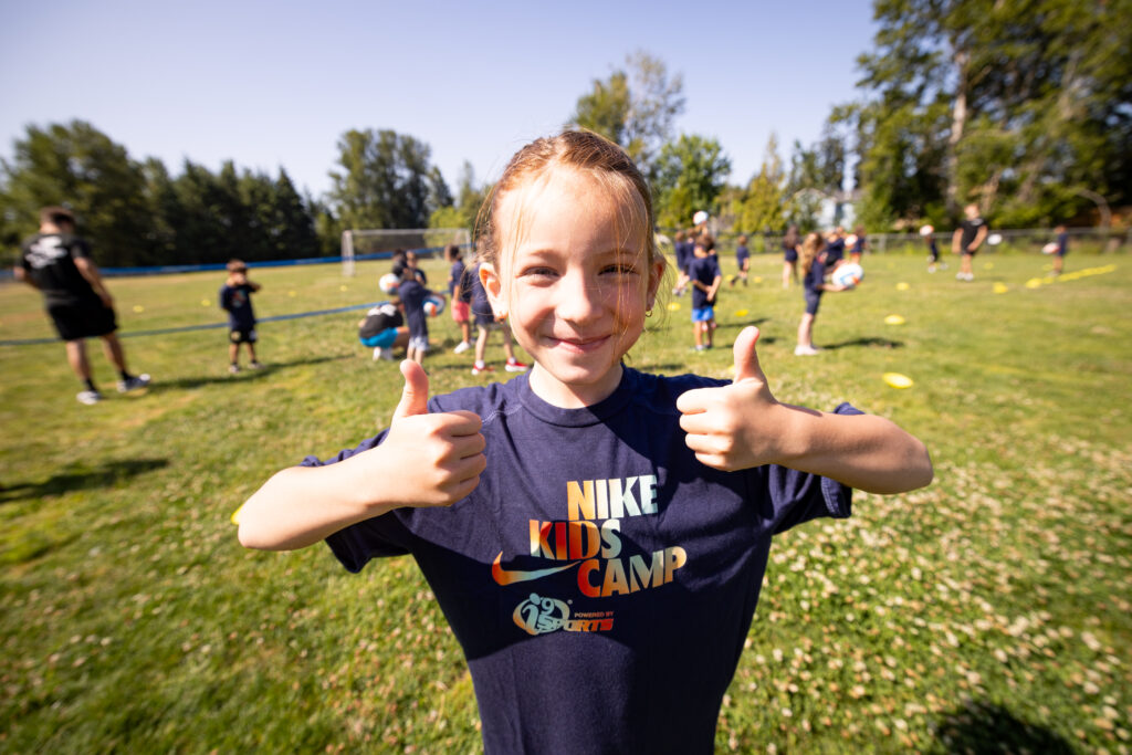 Young blond girl in a dark navy t-shirt that reads "Nike Kids Camp" in orange and green tie dye lettering. She is holding two thumbs up and smiling while learning how to play volleyball.