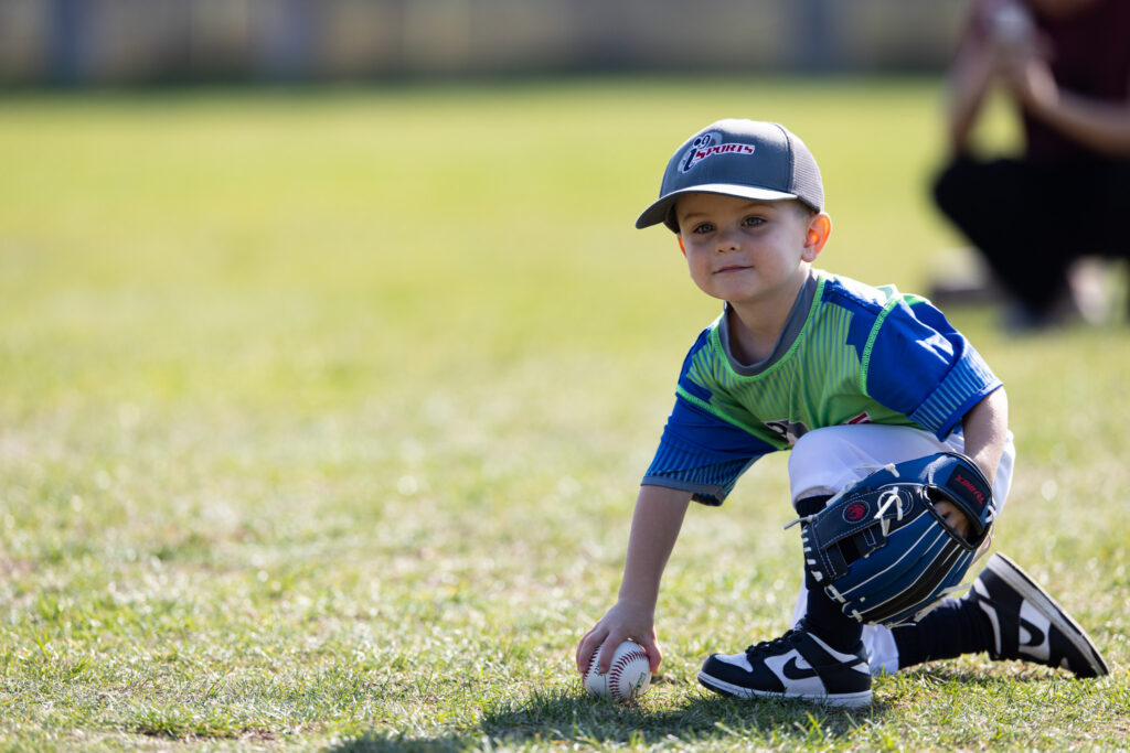 A young boy in his i9 Sports t-ball uniform and a gray i9 Sports baseball cap bending down on one knee to pick up a baseball with his hand.