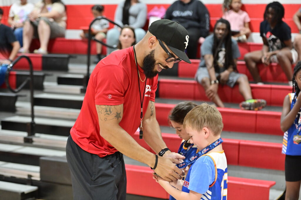 An i9 Sports basketball coach in his red Coach shirt, a black hat, and thick black glasses putting a sportsmanship medal around his young players neck while both he and the player are smiling.