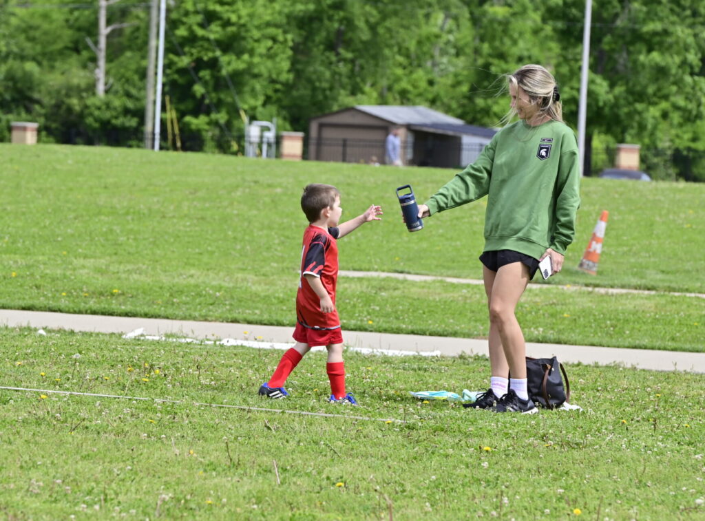 Young soccer player in a red i9 Sports soccer jersey grabbing his navy blue child-sized Yeti water bottle from his mother on the sidelines. Heat safety is important during summer sports!