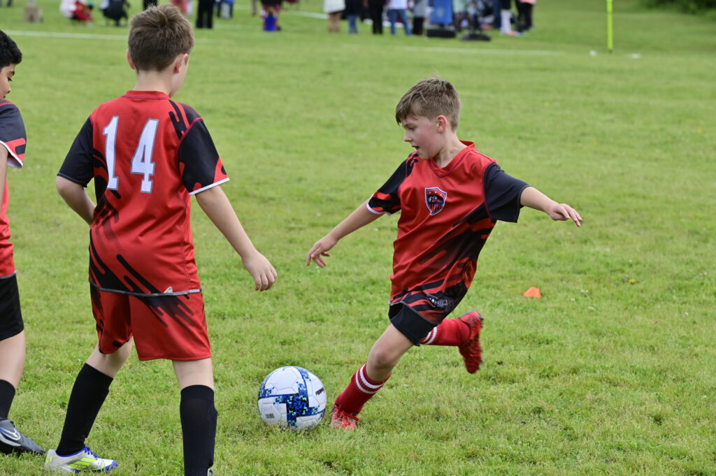 Youth soccer player in his red i9 Sports soccer jersey loading up to kick the soccer ball in front of him.