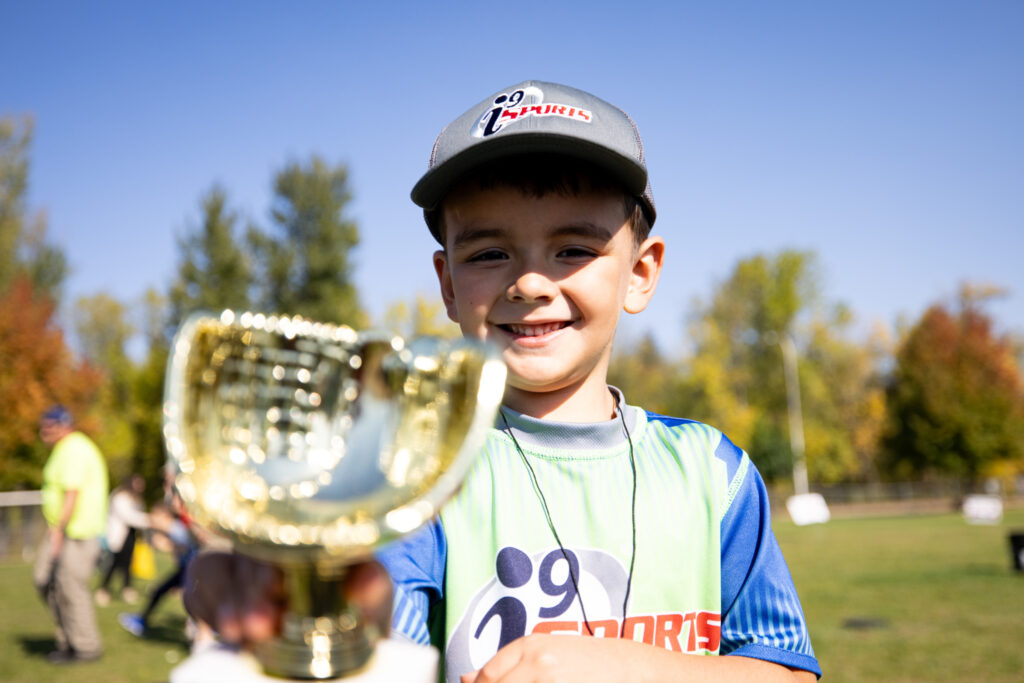 Young boy in a bright green and blue i9 Sports jersey and gray i9 Sports baseball cap holding his baseball glove shaped trophy and smiling.