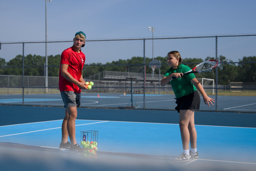 Young beginner tennis coach in a red shirt on the left coaching a pre-teen girl on the right how to swing the tennis racket.