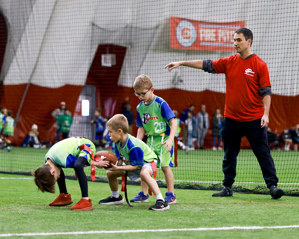 Youth flag football team playing indoor flag football with i9 Sports! The center is snapping the ball to the quarterback while the running back waits behind him. Their coach in a red i9 Sports coach shirt is behind them all pointing to the right (your left).