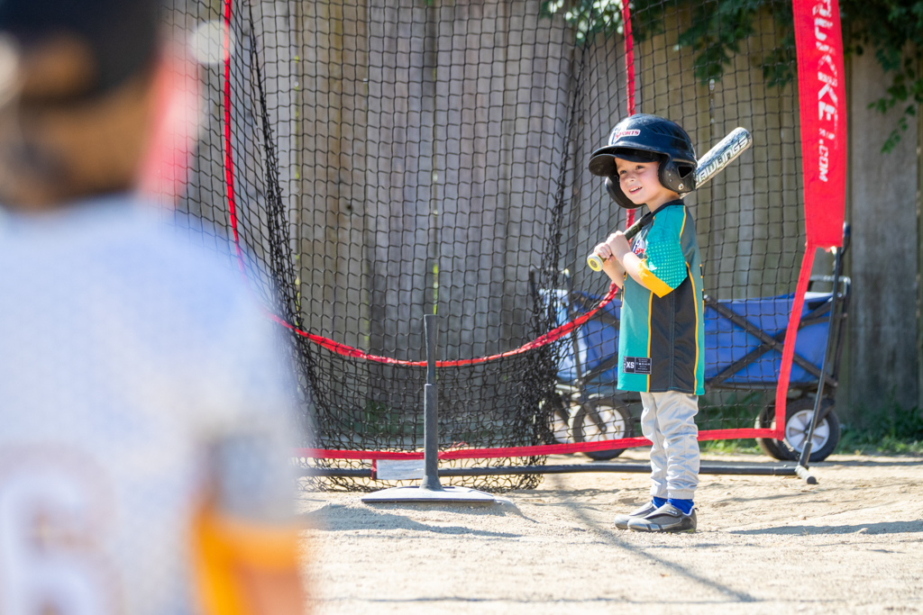 Young boy in his i9 Sports baseball jersey and i9 Sports helmet smiling and standing next to the tee, getting ready to hit during his t-ball game.