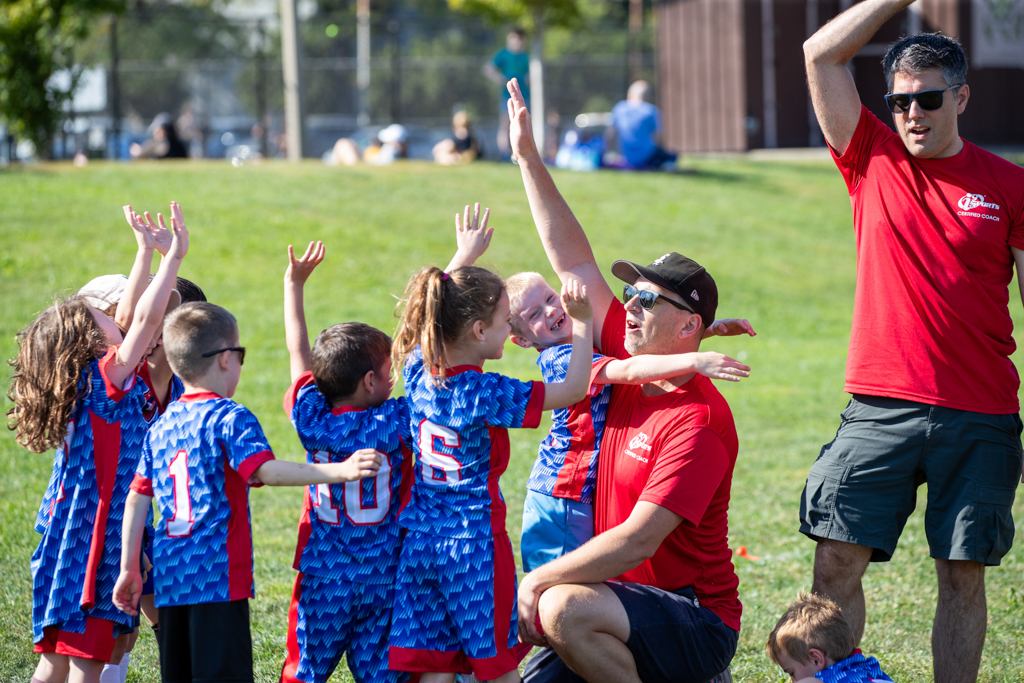 i9 Sports youth soccer team in their bright royal blue jerseys hugging their coach in his read i9 Sports coach shirt. Skipping the travel team allows your kids to have fun and grow at their own pace, making them more likely to stick with sports longer.