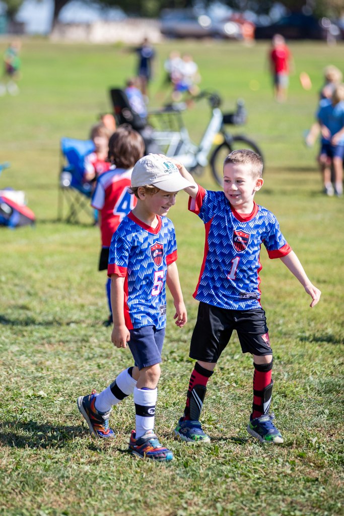 Two young boys smiling while walking down the soccer field. The boy on the right is tapping the boy on the left on the head while the boy on the left laughs.