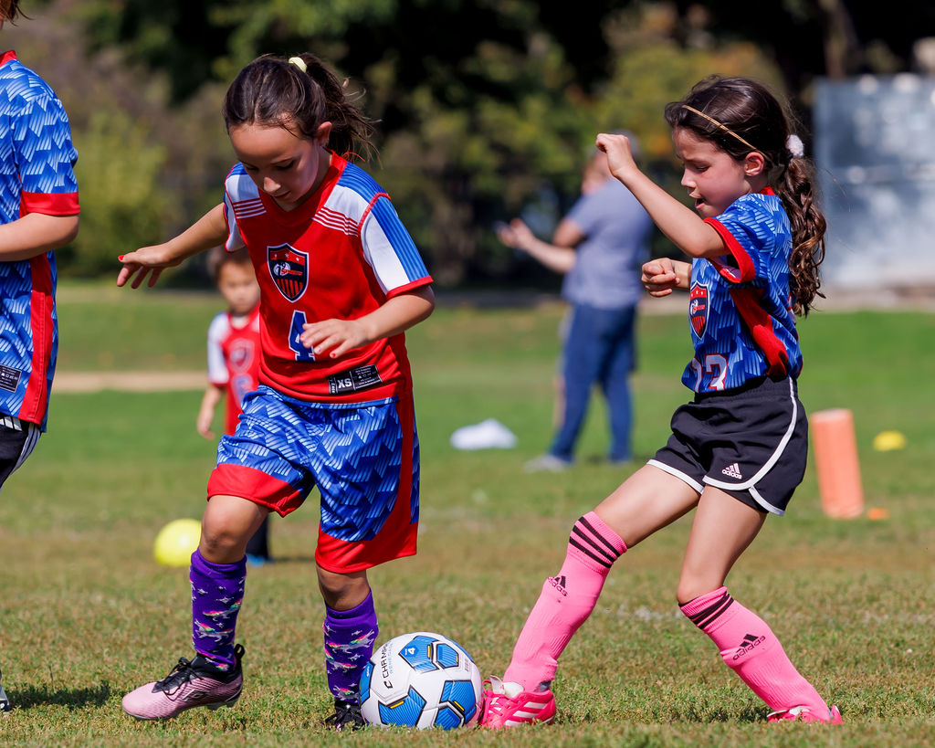 Two girls playing soccer with i9 Sports. Girl on the right in a blue jersey is kicking the ball with her pink socks and pink cleats. While girl on the left in the red jersey is trying to defend.